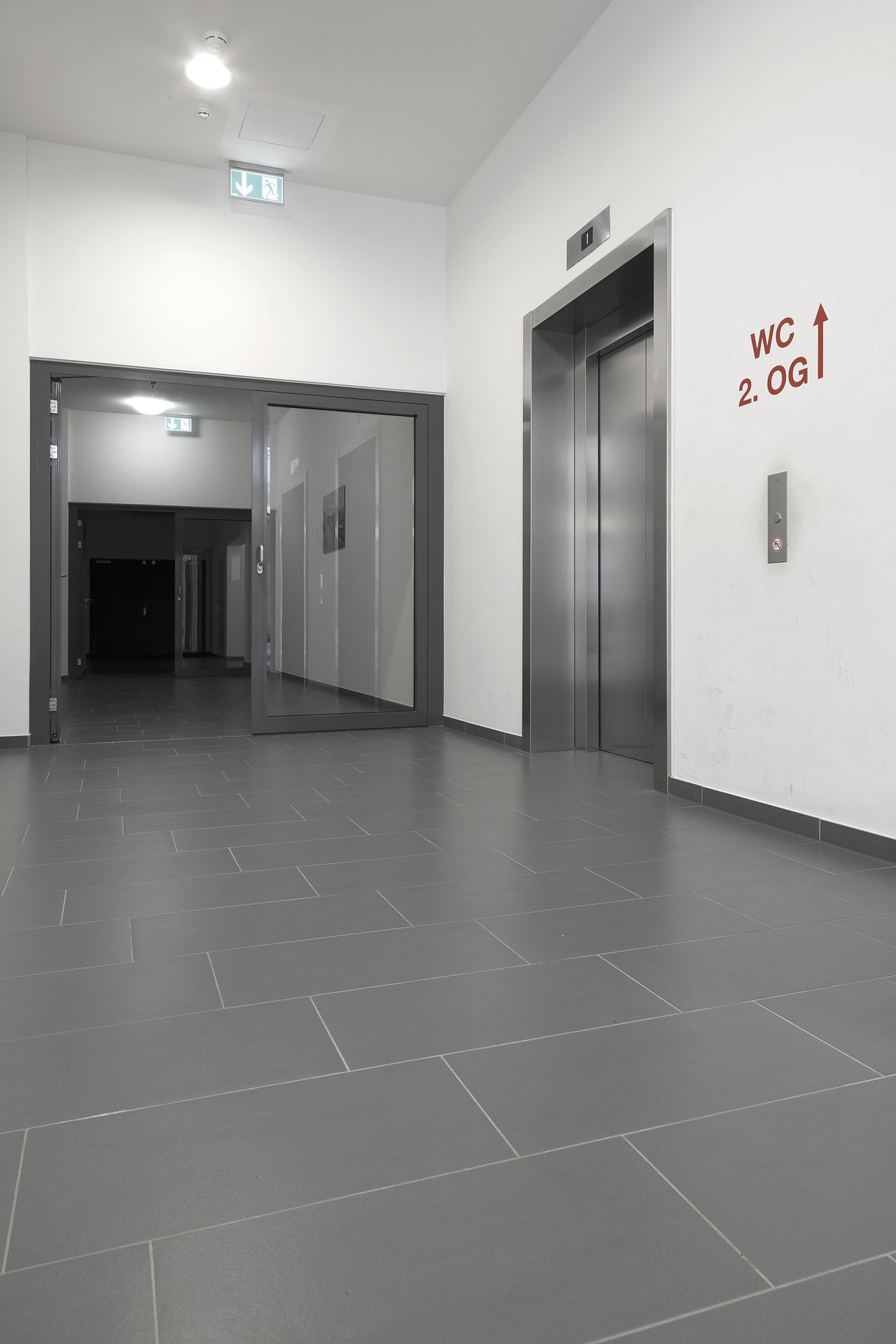 Modern hallway with large-format grey floor tiles and elevator.