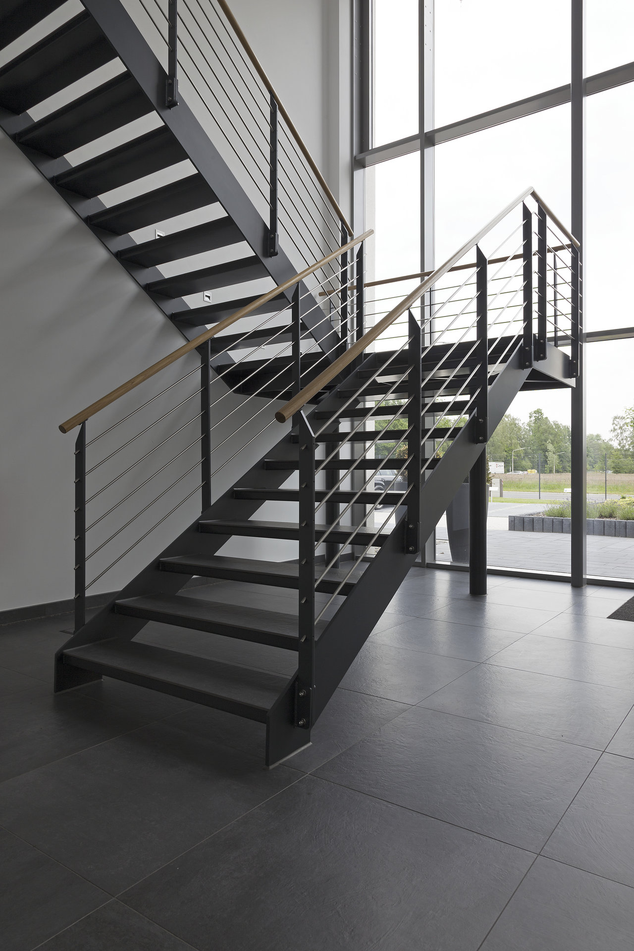 Modern dark tiles in the entryway under a steel staircase with a wooden handrail.
