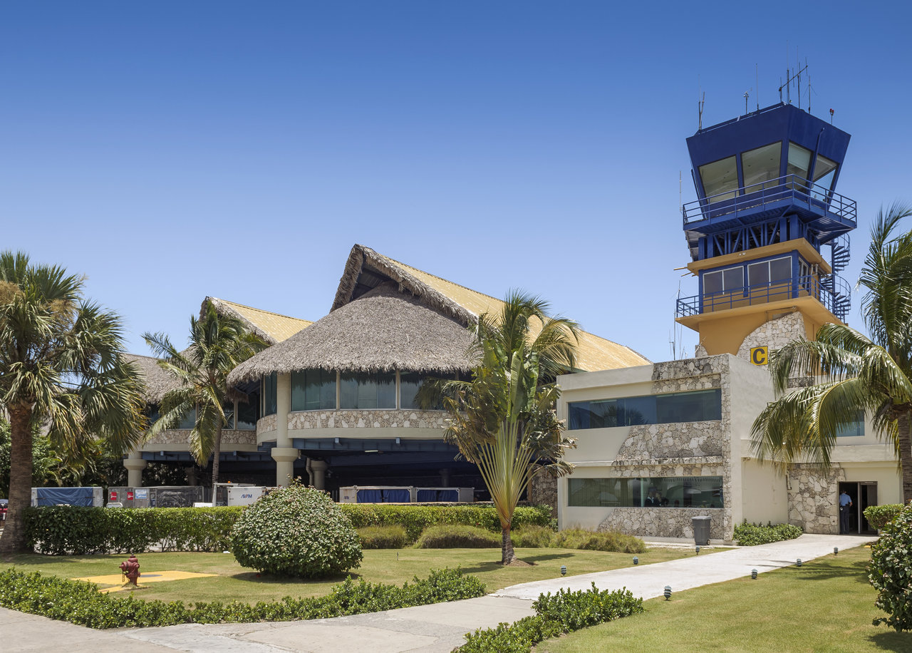 Tropical airport with thatched-roof building, palm trees, and blue-yellow control tower.