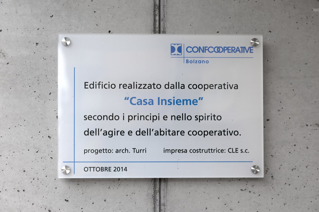 White plaque on concrete wall, detailing cooperative building project in Bolzano.