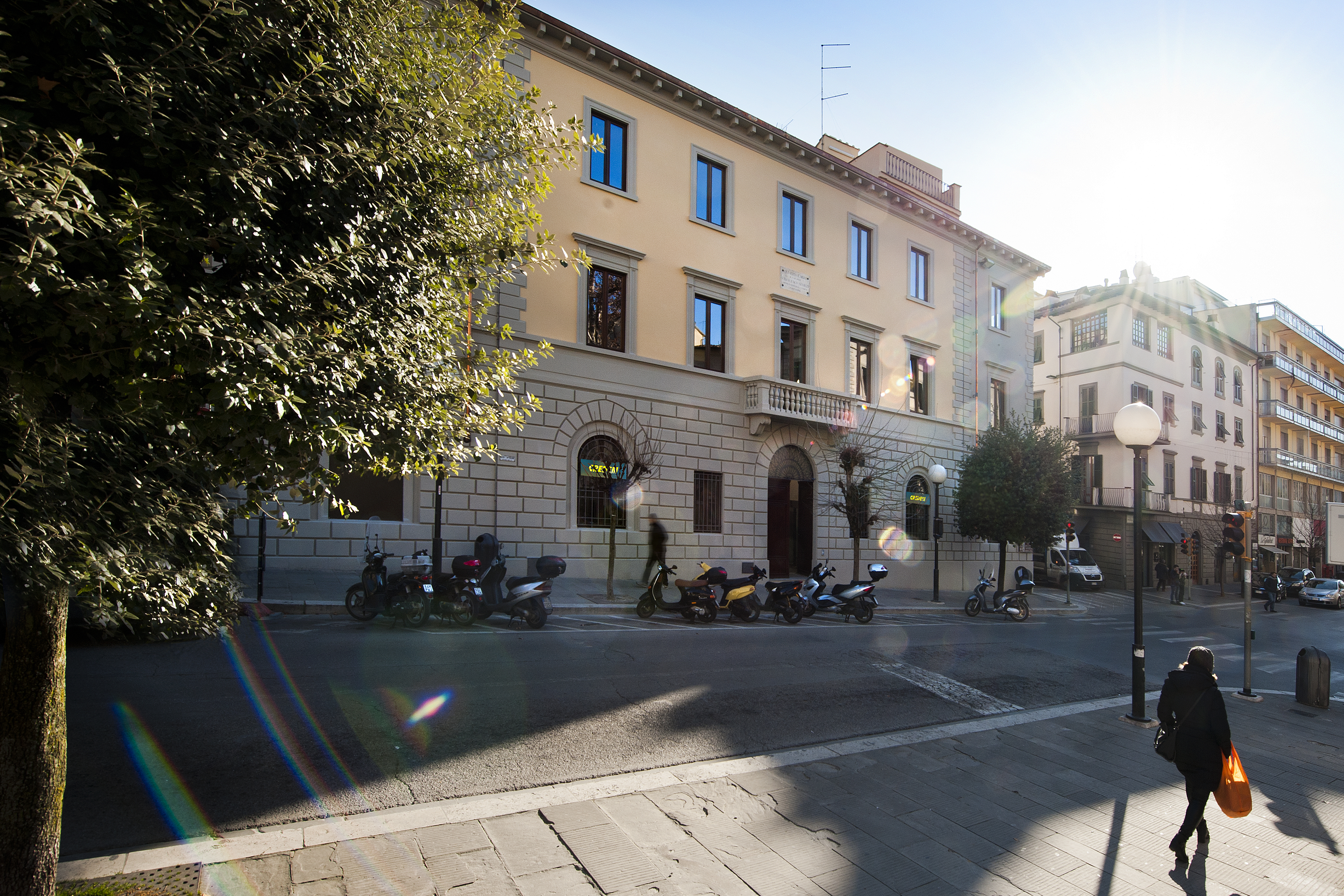 Sunny city view with elegant building, parked scooters, and a person on the pavement.