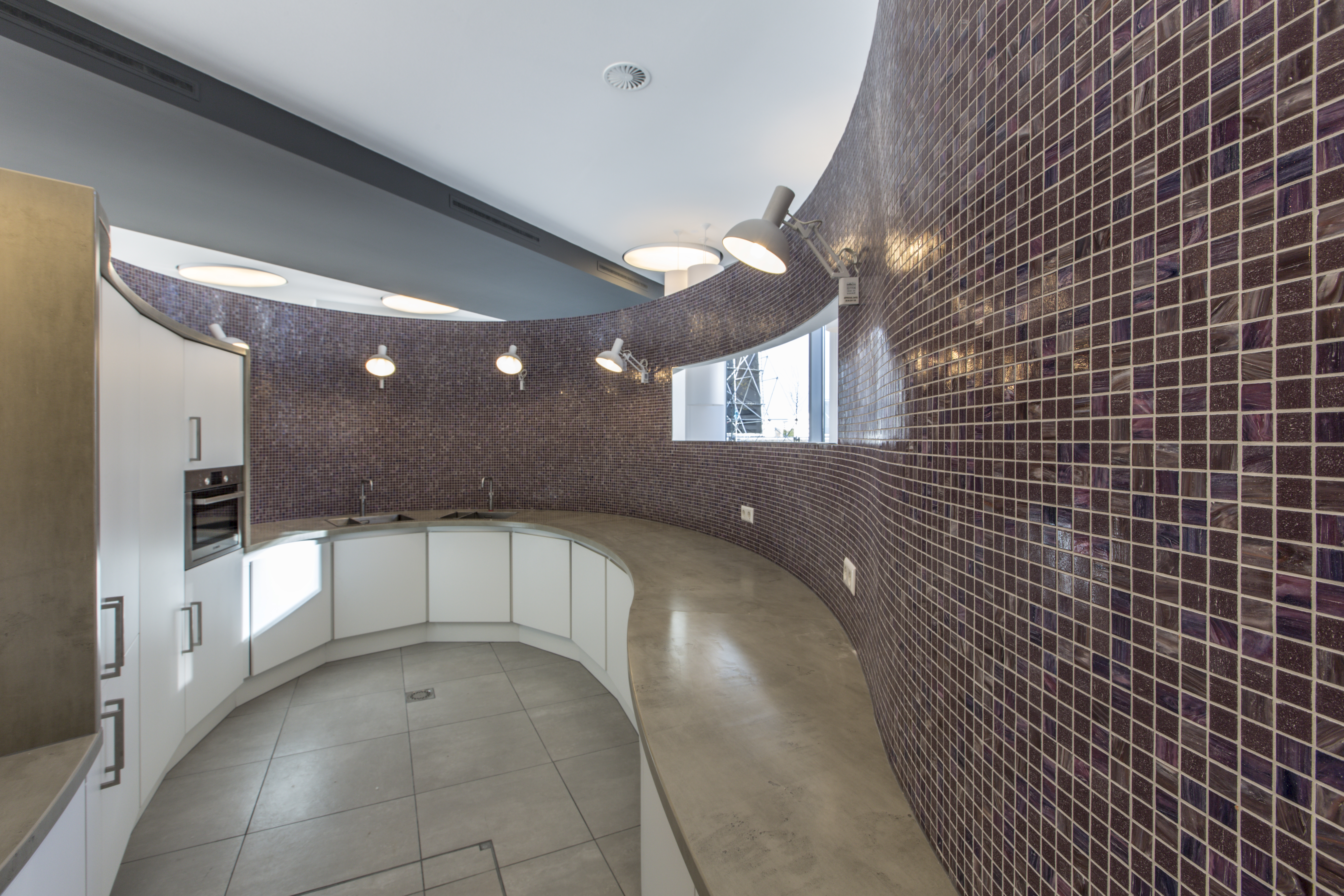 Modern kitchen with a curved mosaic tile wall, concrete-look counter, and light tiled floor.