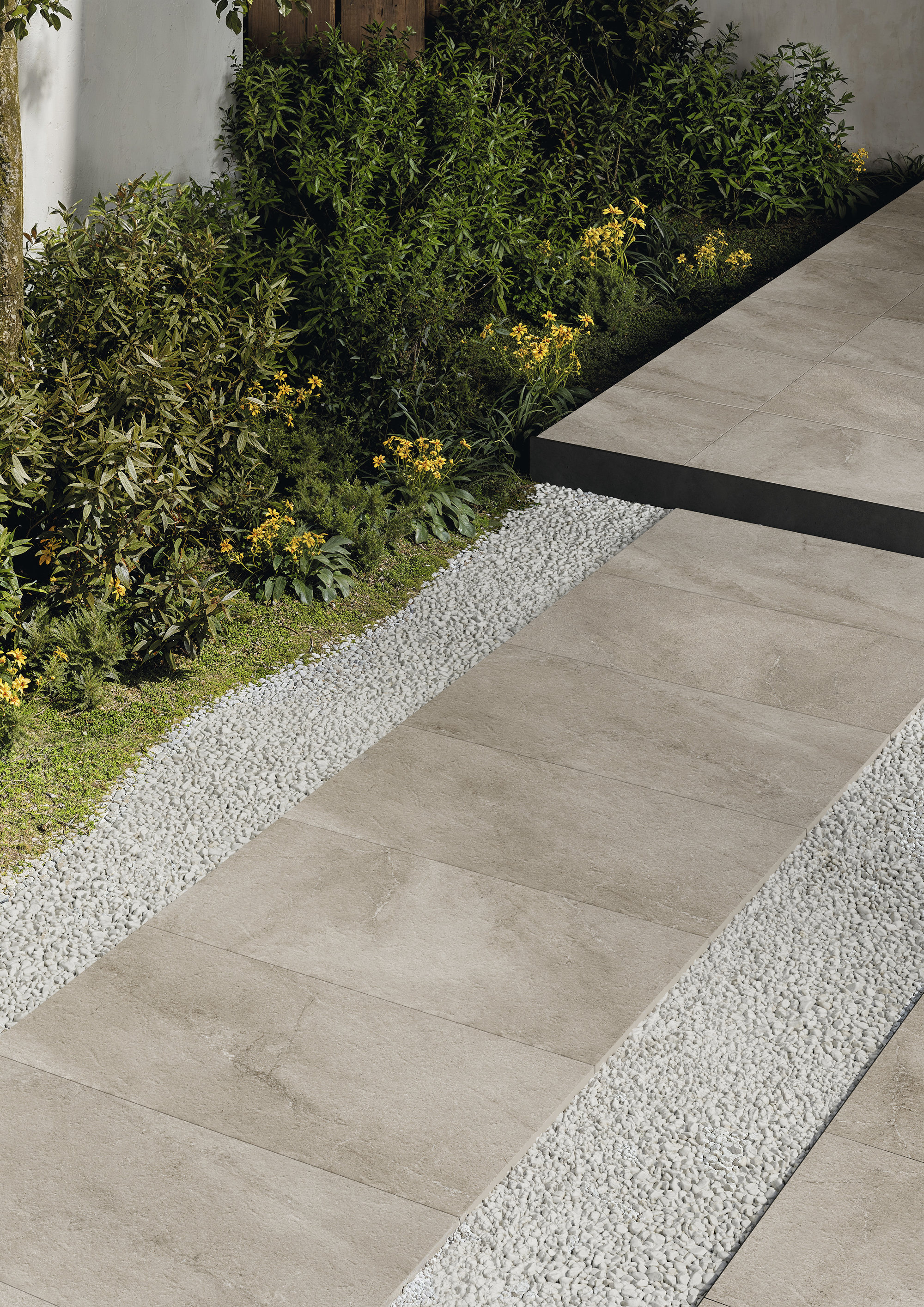 Modern grey outdoor tiles as a path and raised area, surrounded by gravel and green plants.