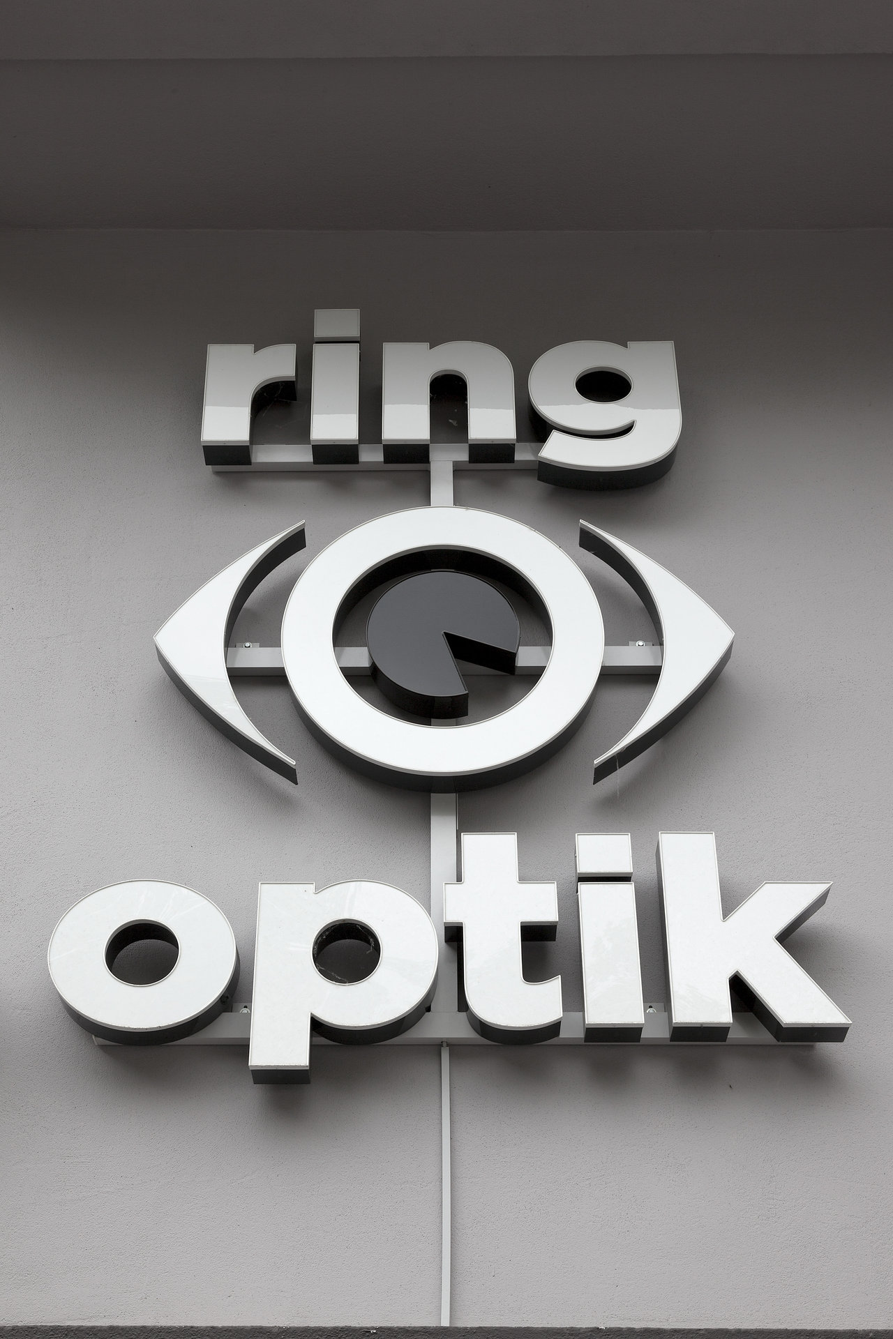 Ring Optik's silver 3D sign with stylised eye on a grey wall.