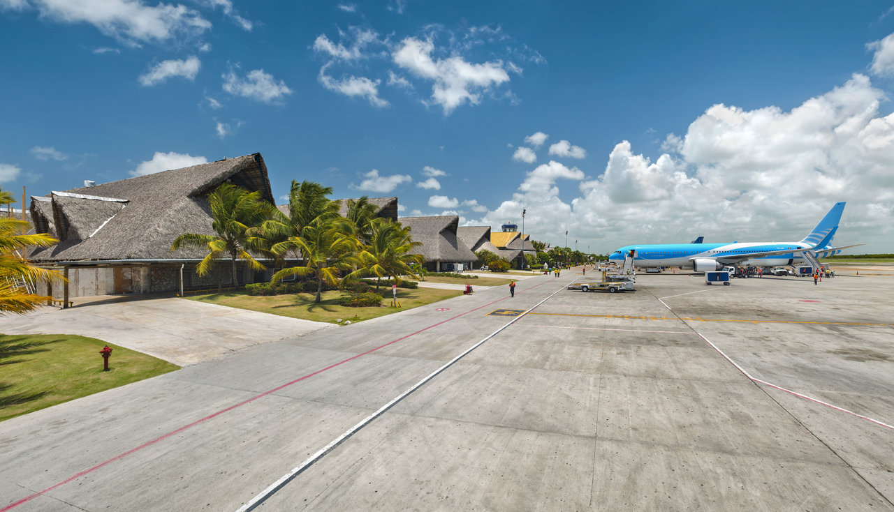 Airport in a tropical landscape with palm trees, thatched-roof buildings, and a blue plane.