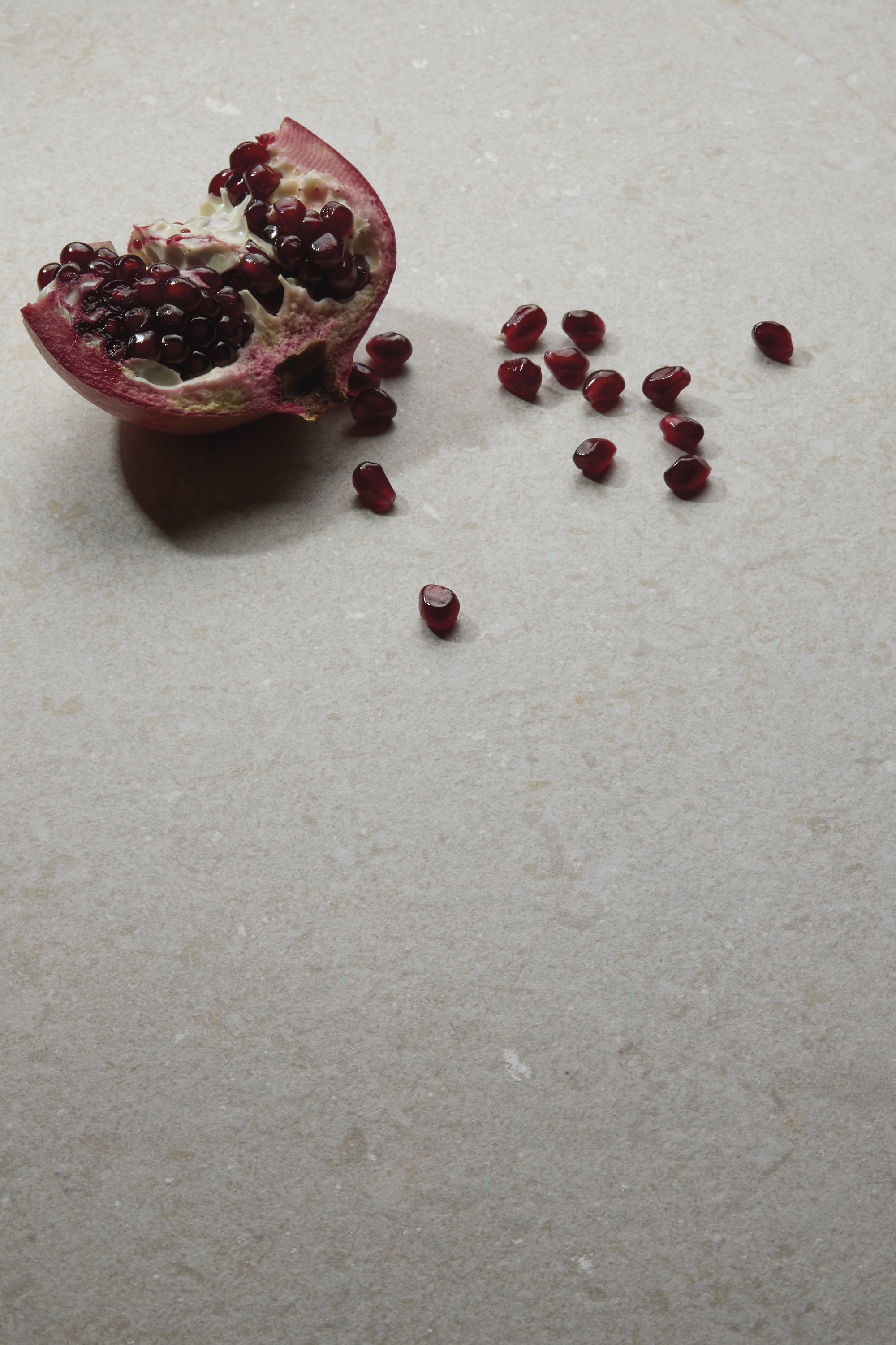 Pomegranate half and individual seeds on light grey, textured tile.