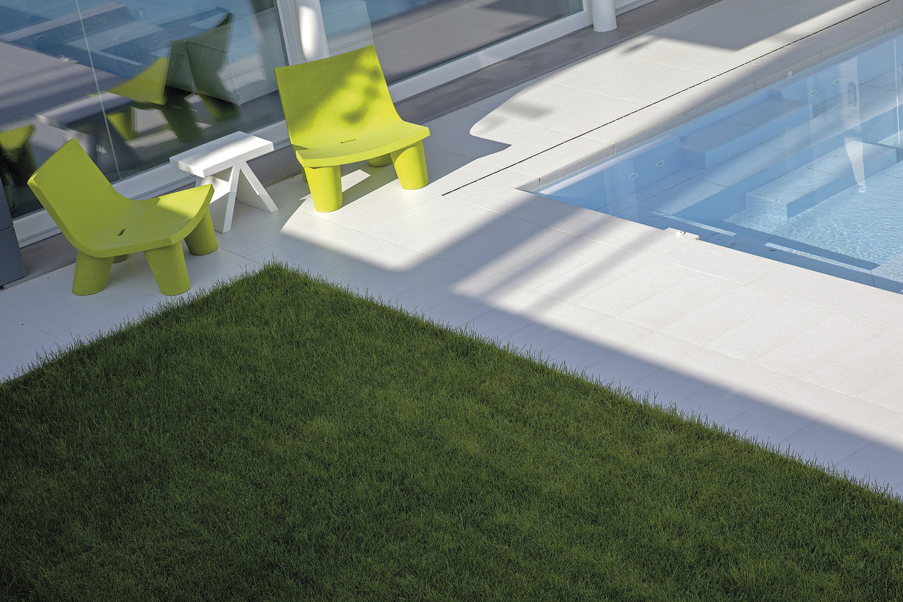 Modern pool surround with light tiles, lawn, and two green chairs.
