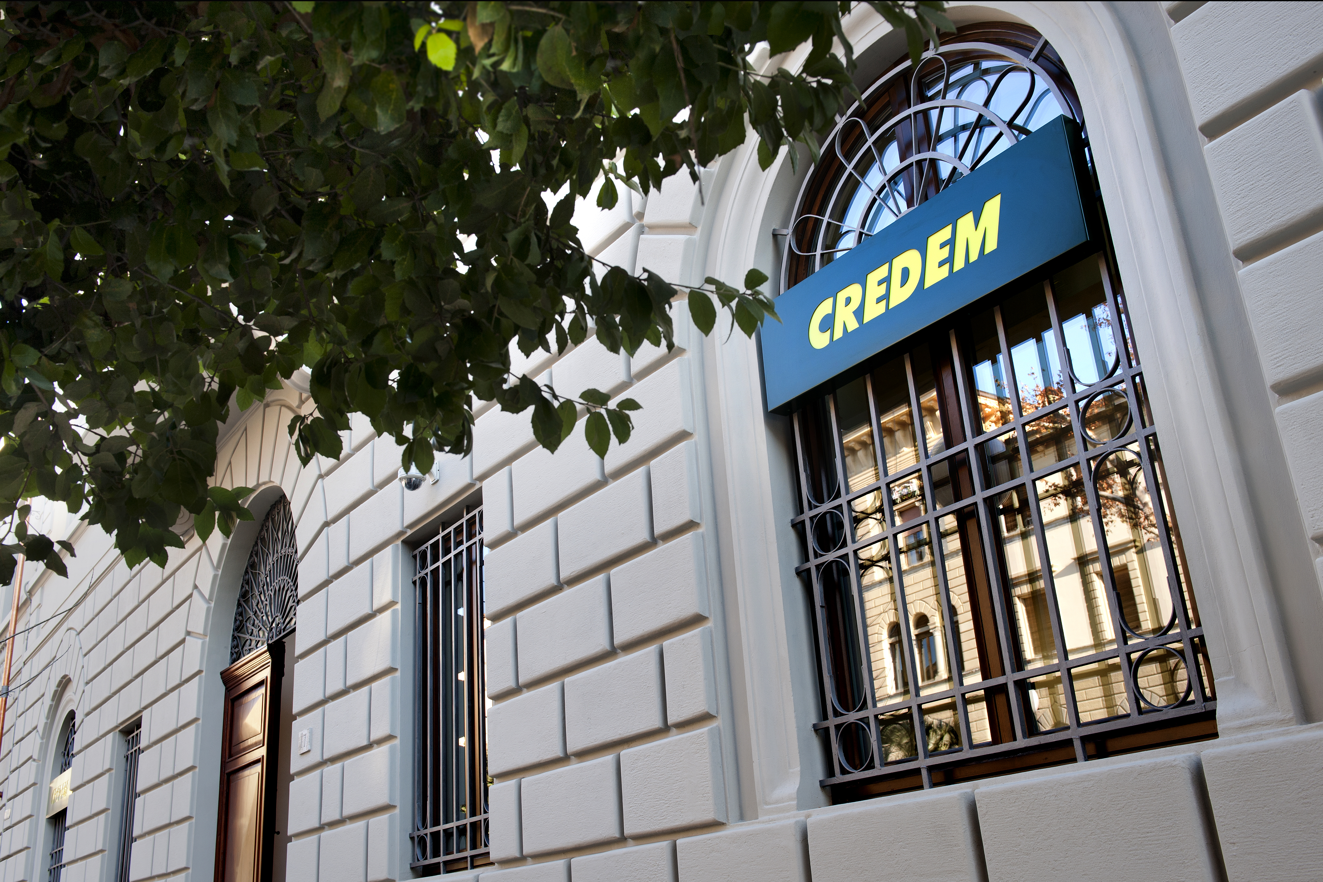 Building facade with 'CREDEM' on window, tree leaves in foreground.