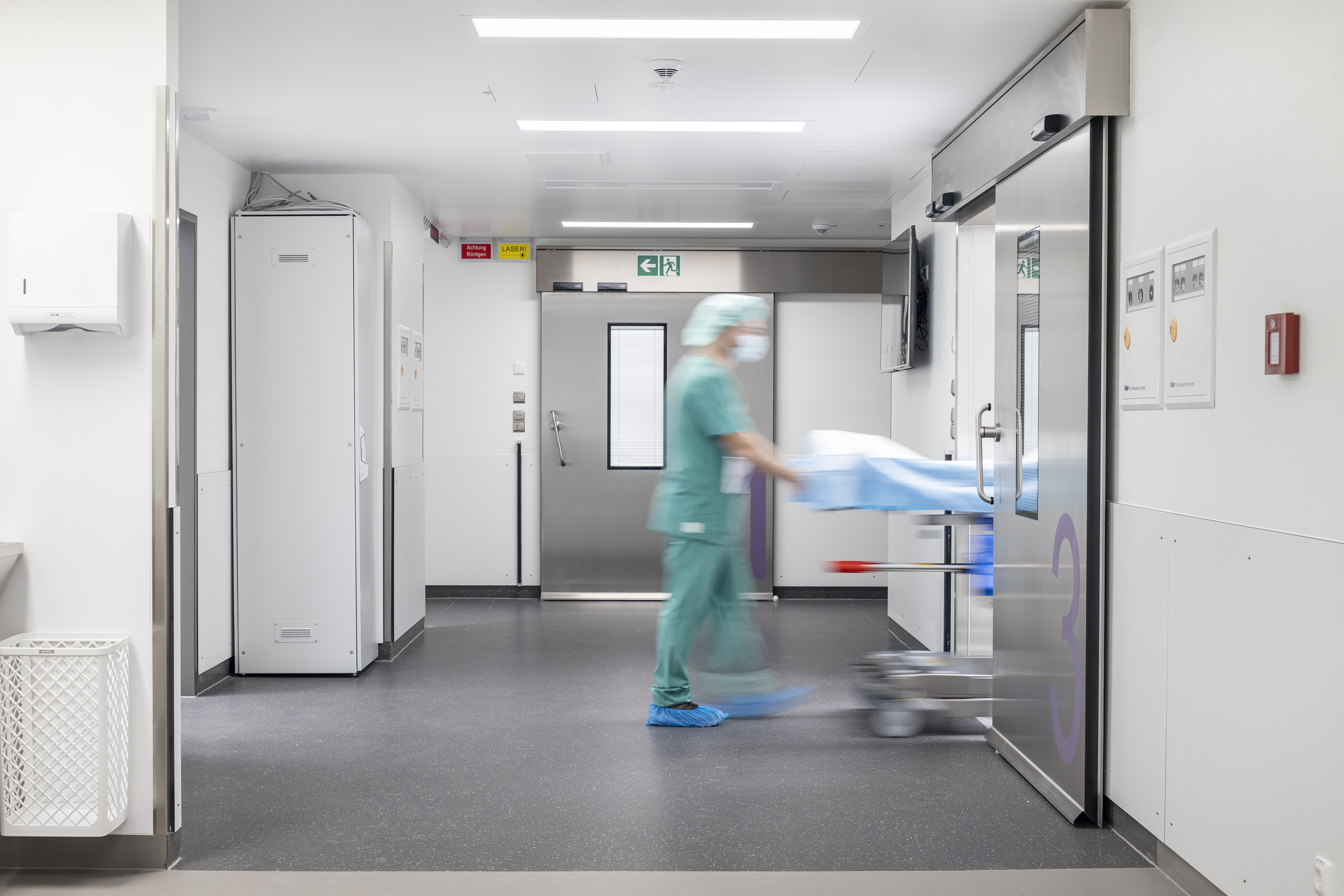 Modern hospital corridor with dark floor tiles and a rushing person with a stretcher.